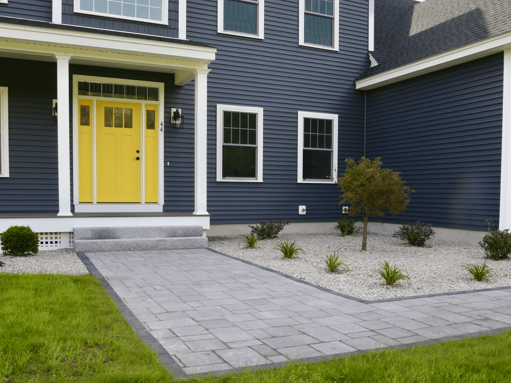 Gray paver walkway and stone landscaping in front of a blue house with a yellow front door.