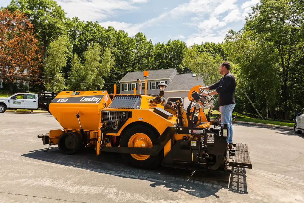 Orange asphalt compactor/road roller in a parking lot, operator standing on the platform with a suburban neighborhood in the background.