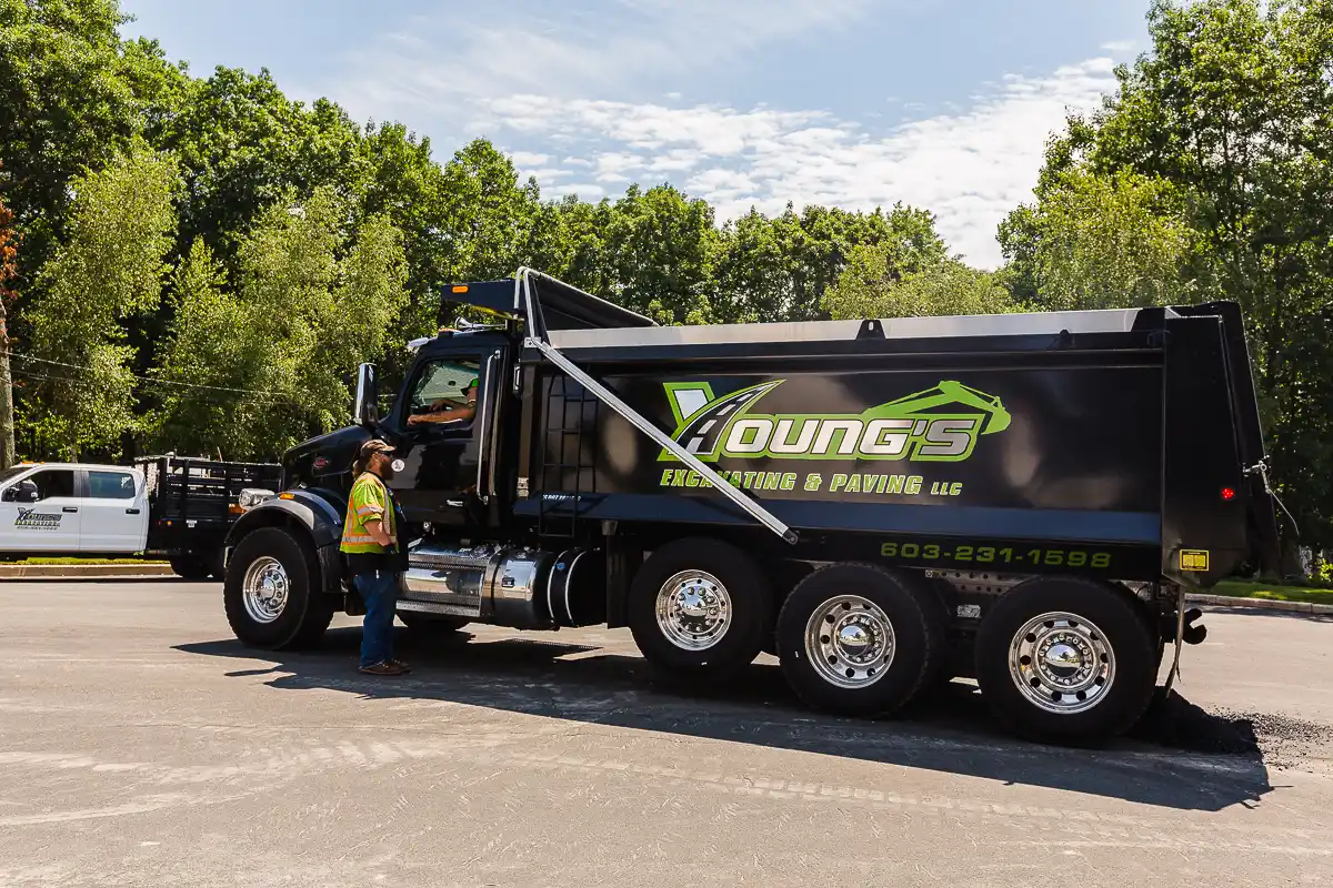 Large black dump truck with green logo reading 'Young's Excavating & Paving' parked in a lot, a worker in a safety vest nearby inspecting the cab.