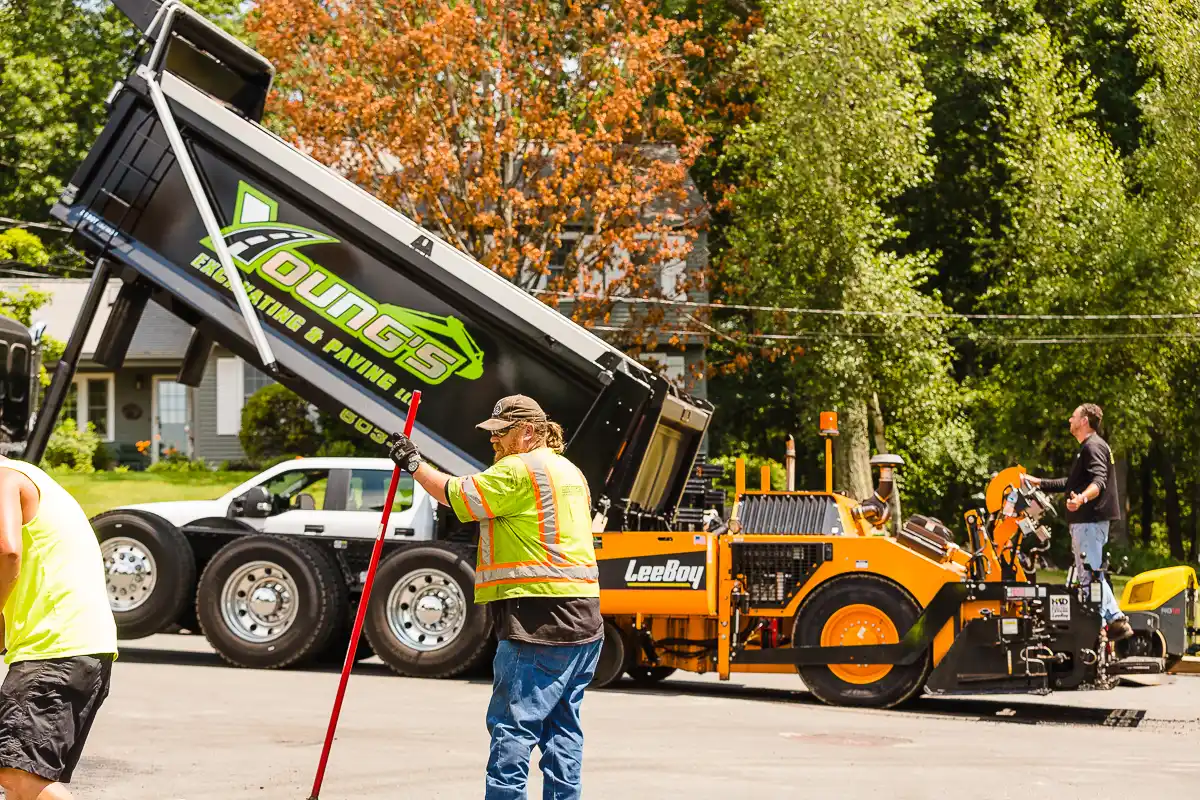 Young's Excavating & Paving LLC dump truck unloading hot asphalt into a paver while the crew directs, Penacook, NH.