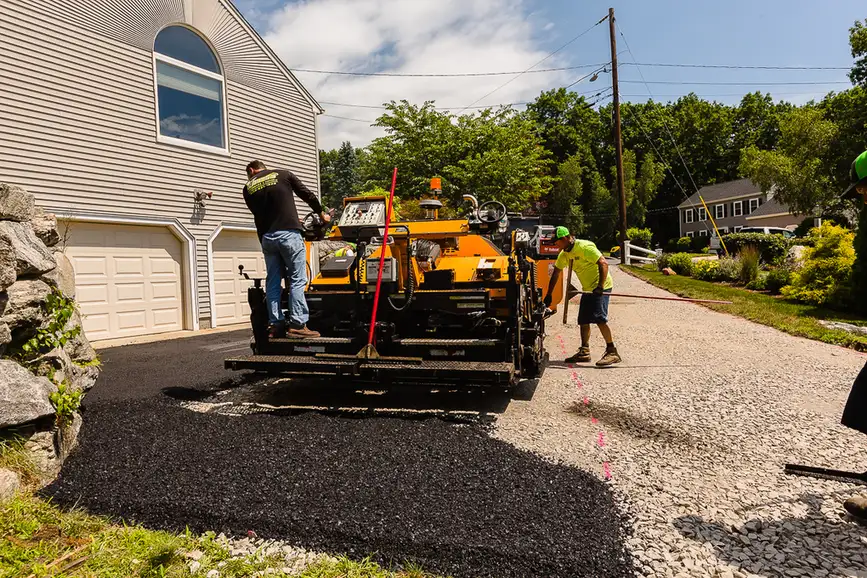 Two workers operate a yellow asphalt paver laying fresh blacktop on a driveway in front of a house.