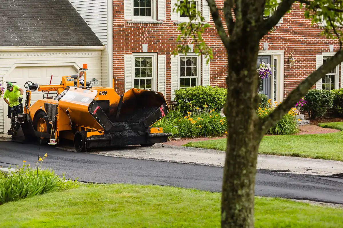 Orange asphalt paver laying a new road surface in front of brick houses, with a worker nearby by the driveway.