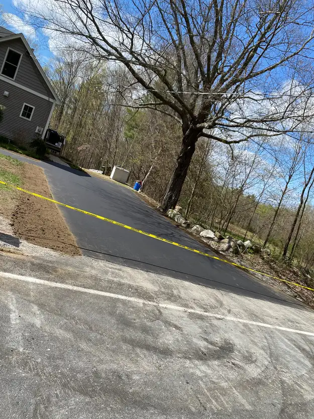 Freshly paved asphalt driveway cordoned off with yellow tape beside a house and a large tree in a wooded yard.