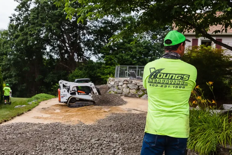 Track loader spreading crushed stone base on a residential driveway while a Young's Excavating & Paving LLC crew member watches.