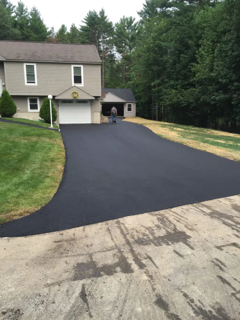 A newly paved black asphalt driveway curves up to a beige two-story house with a white garage door; a man walks toward the garage.