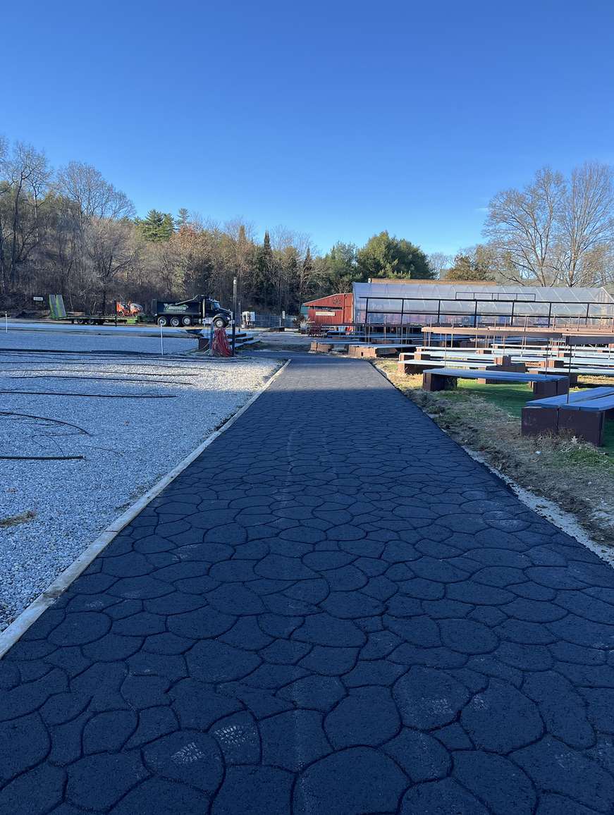 Wide view of an outdoor recreation area: a cracked asphalt path leads toward a greenhouse and red shed, with benches and a gravel lot nearby under a clear blue sky.