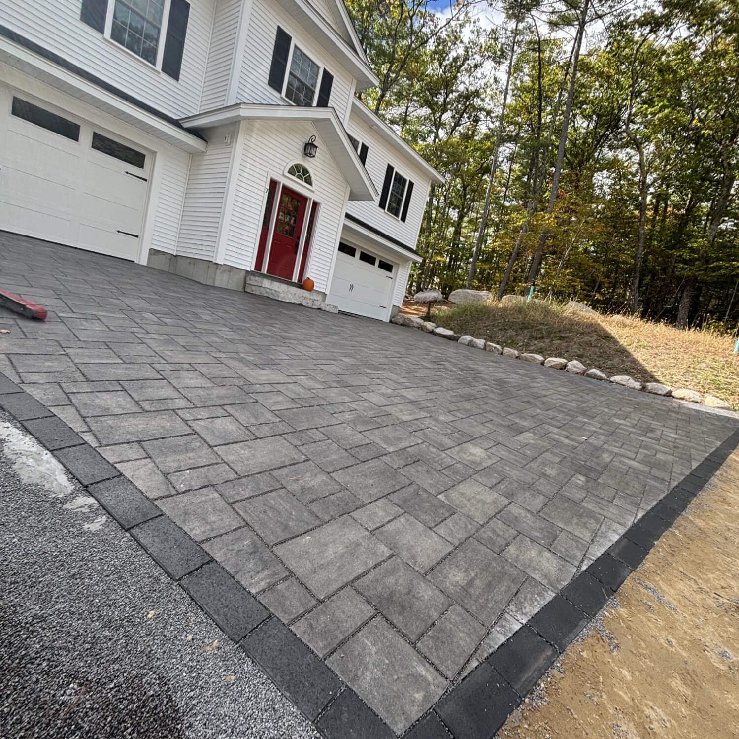 Gray paver driveway with dark border in front of a white house surrounded by trees.