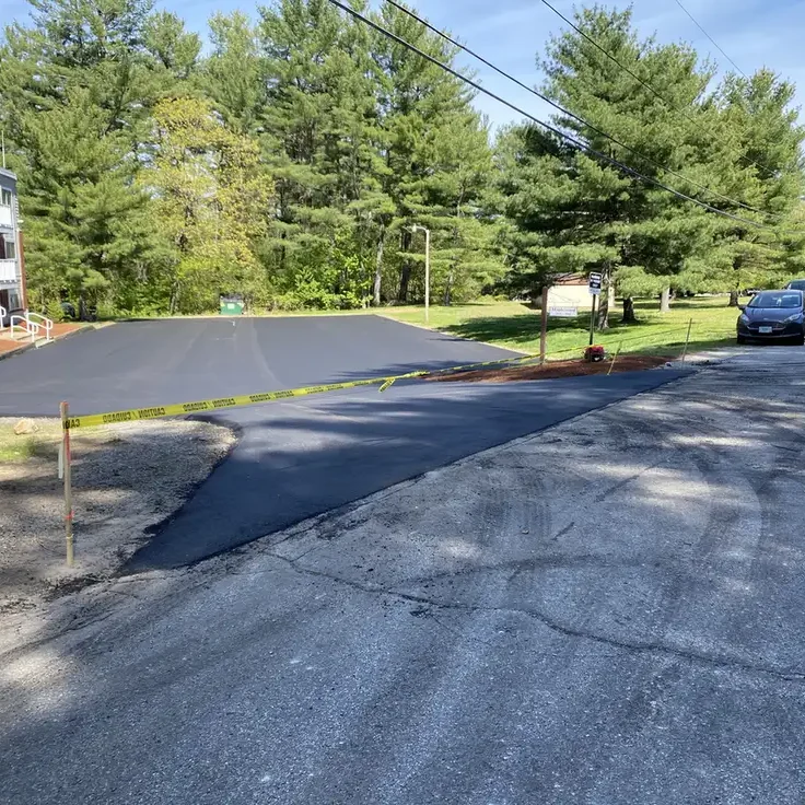 driveway paving londonderry nh, new asphalt patch being laid on a residential street, with yellow caution tape, parked cars to the right and an apartment building to the left.