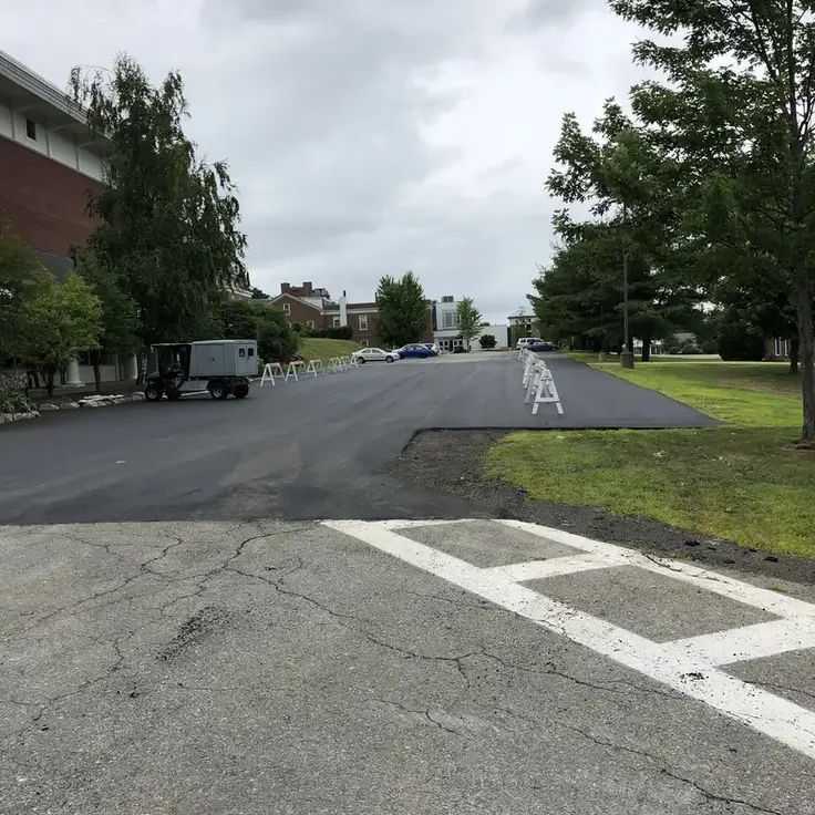 Parking lot under construction with fresh asphalt, white barricades, and a small truck on the left; overcast sky and trees along the sides.