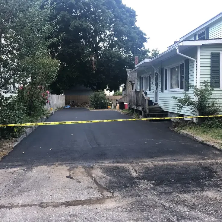 A residential driveway blocked by yellow caution tape, leading to a paved path between houses with trees and a sign on the right.