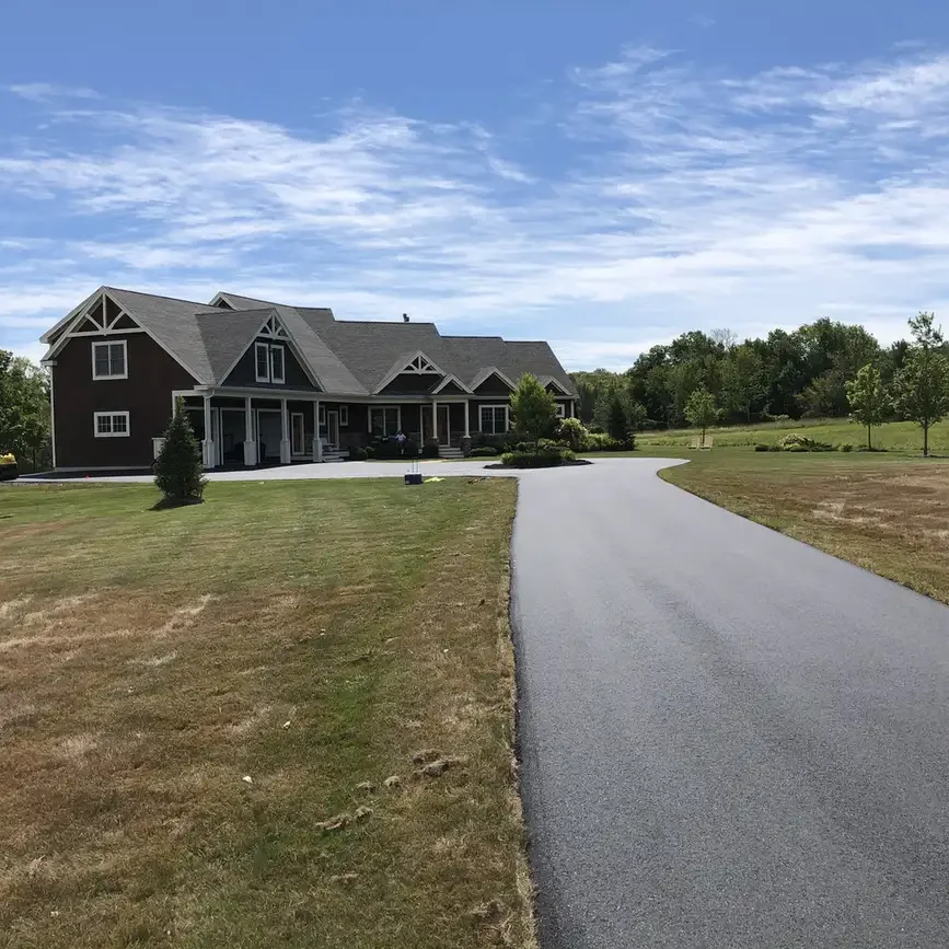 A large dark-brick house with a gray roof sits at the end of a curved asphalt driveway on a spacious lawn under a blue, partly cloudy sky.