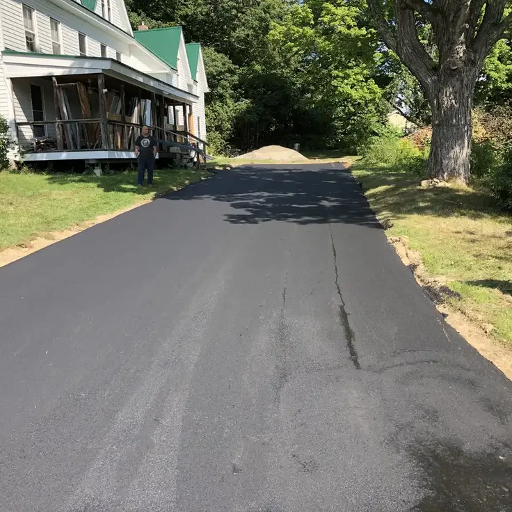 New asphalt road under construction in a residential area, a man stands near a white house with a wraparound porch on the left; a dirt pile sits at the road's end under trees on the right.