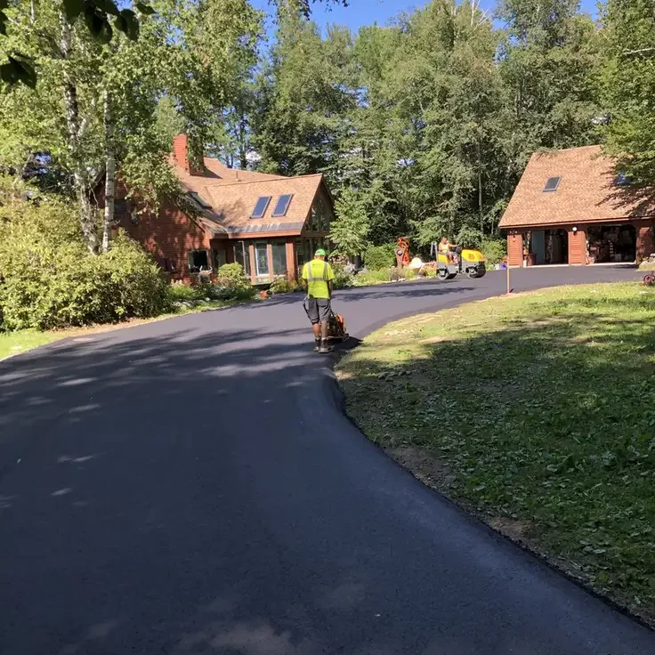 A worker in a high-visibility vest paves a new asphalt road in a residential neighborhood with wooden houses and green trees nearby.