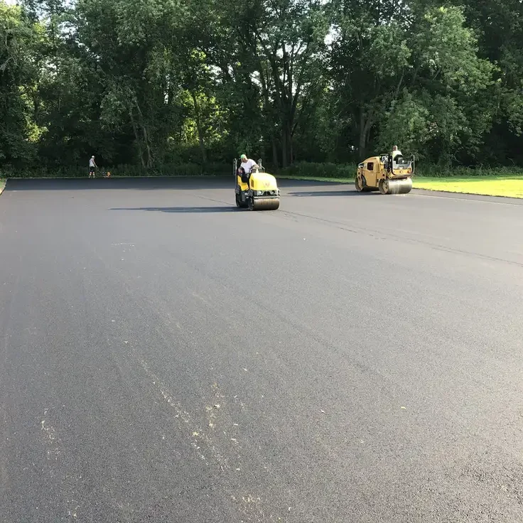 Two yellow road rollers compacting freshly laid asphalt on a wide, empty road with workers and green trees in the background.