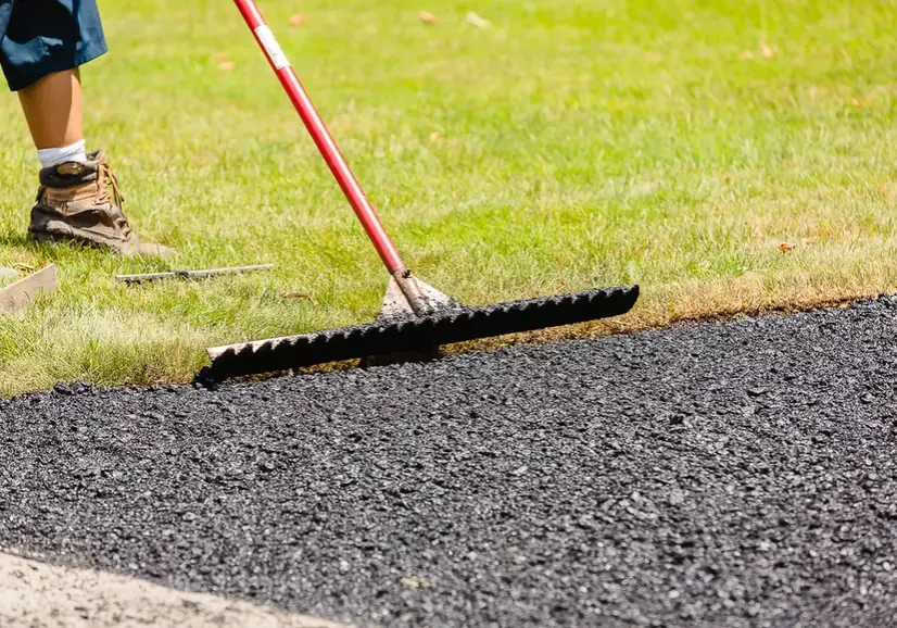 Close-up of a red-handled rake smoothing fresh asphalt along a grassy edge, with a worker's boot visible in the background.