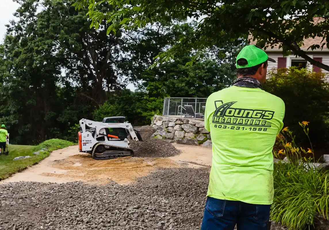 Crushed stone base preparation. Crew member and compact skid-steer loader during residential site work, excavated rock piled nearby.