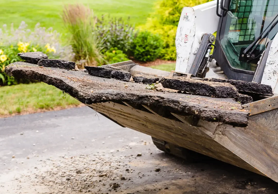 Skid-steer bucket holding broken-out asphalt during driveway removal and excavation.