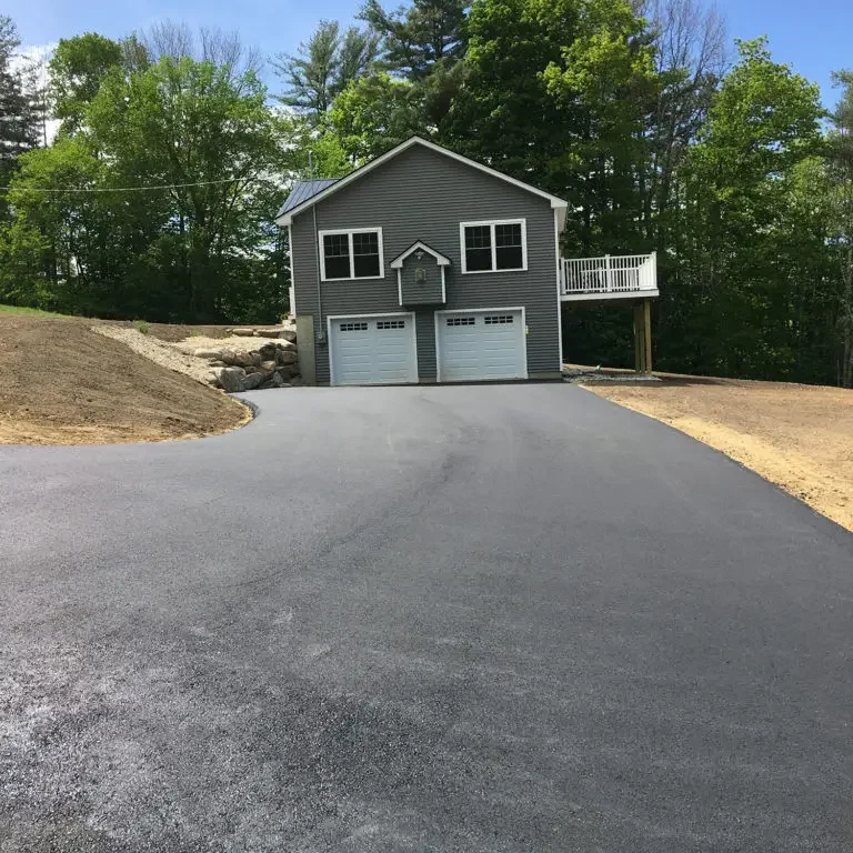 Gray two‑story house with a double garage at the end of a freshly paved driveway, surrounded by trees on a sunny day.