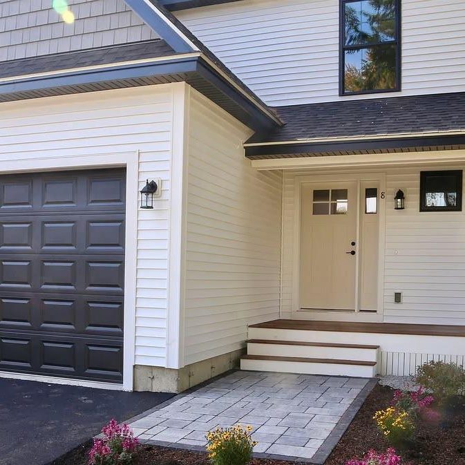 Front entry of a modern home with a beige door, small window, and porch steps; white siding and a two-car dark gray garage door to the left.