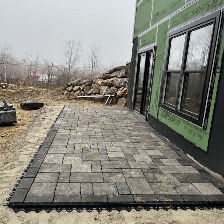 Newly constructed house with green siding sheathing and a rectangular gray concrete-paver patio along the side.