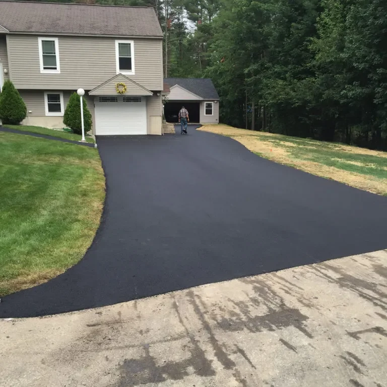 A newly paved black asphalt driveway curves up to a beige two-story house with a white garage door; a man walks toward the garage.