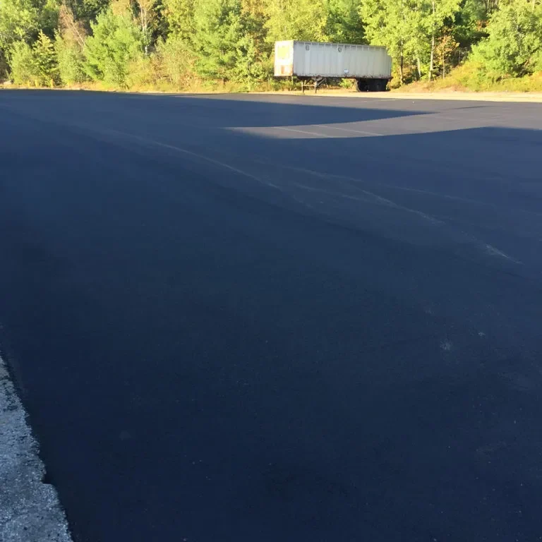 Freshly paved blacktop lot with a white trailer parked near the tree line in the distance.
