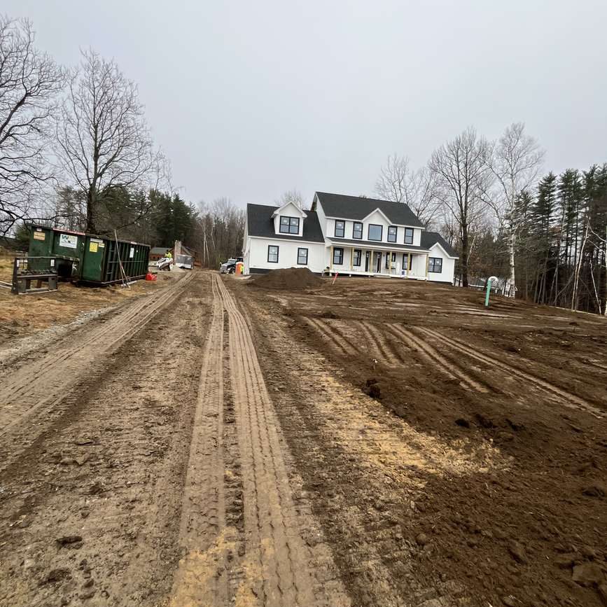New two-story white house under construction at end of a dirt road; muddy path with tire tracks, dumpsters on the left and bare trees in the background.