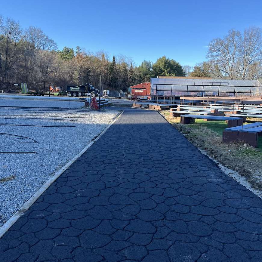 Wide view of an outdoor recreation area: a cracked asphalt path leads toward a greenhouse and red shed, with benches and a gravel lot nearby under a clear blue sky.